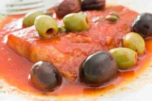 primo piano di una fetta di pesce spada alla livornese con olive e capperi e sugo di pomodoro